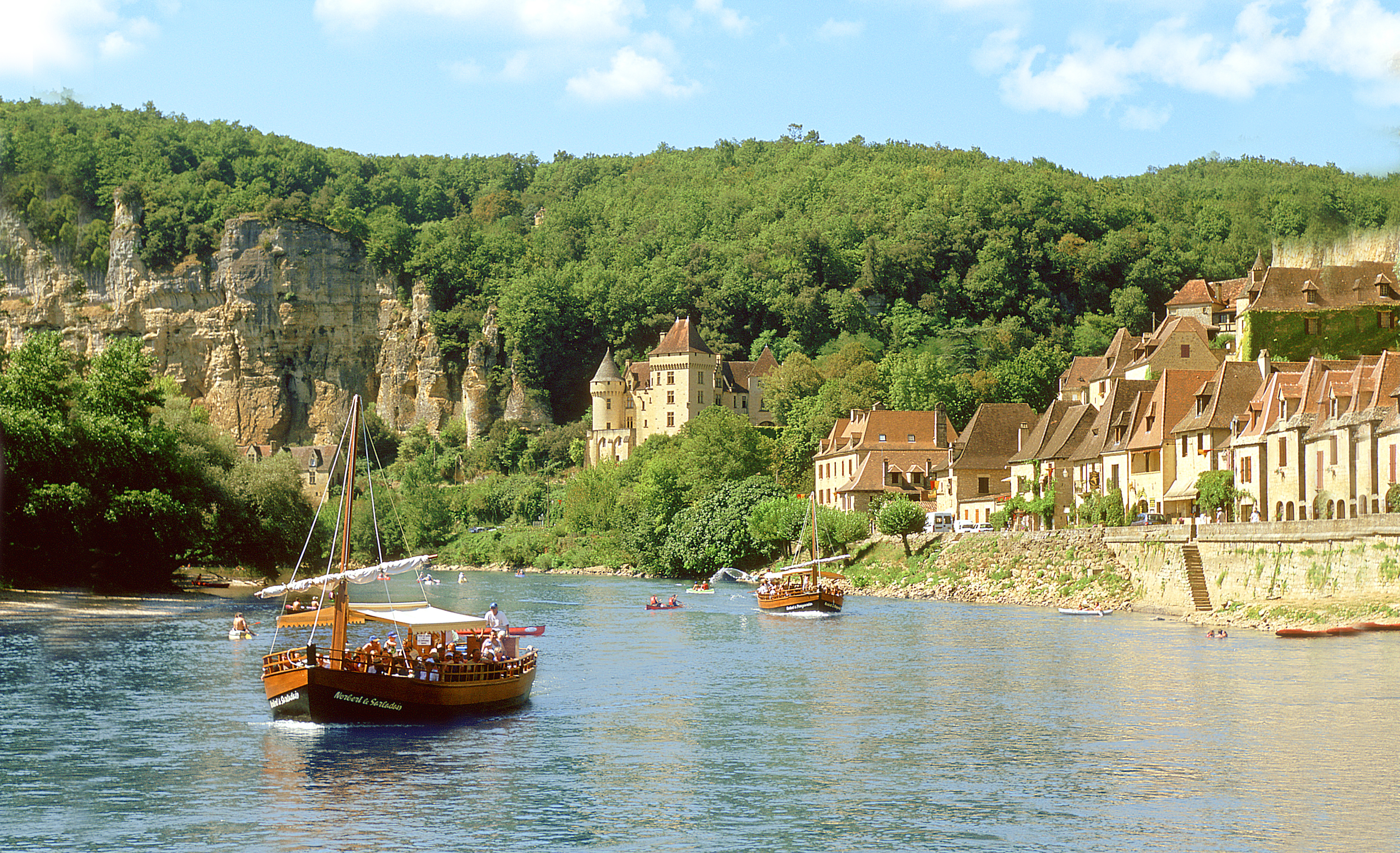 Two boats in front of a hillside village full of greenery, with people in kayaks in the water as well.