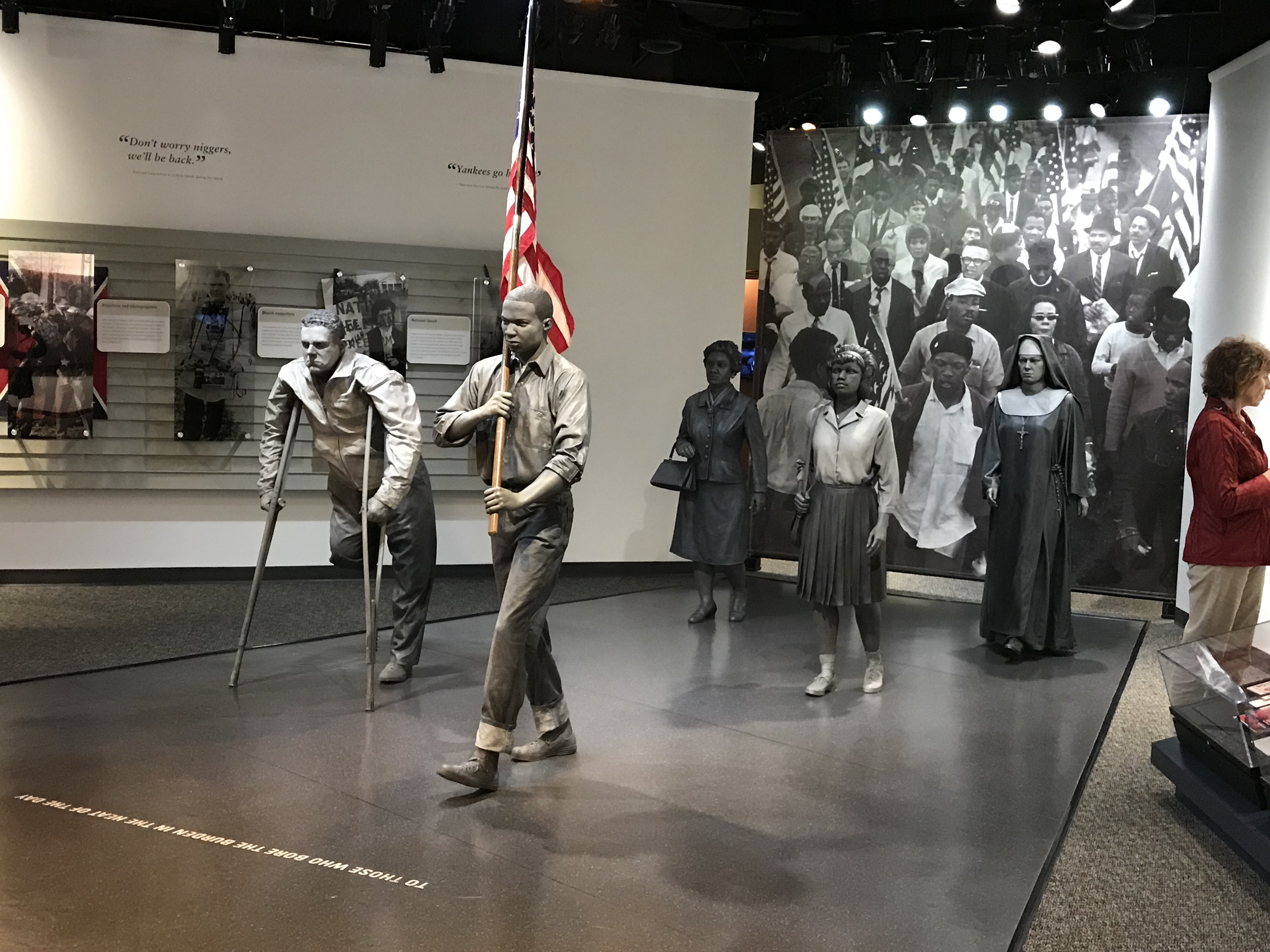 Inside the Lowndes Interpretive Center. Grey tone sculptures of men and women, one holding an American flag, in the center of the room, with images and text on the walls. 