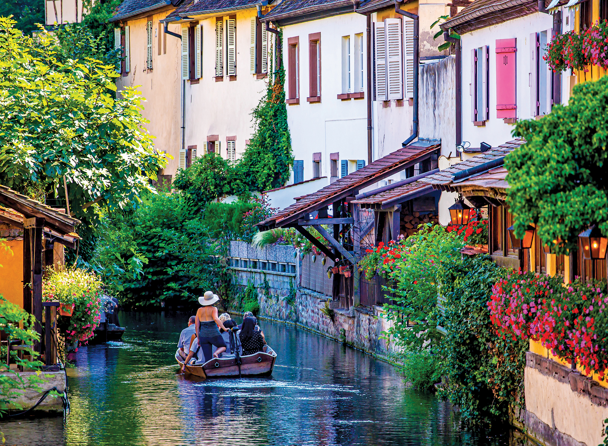 A small boat with a few people travels down a river, away from the viewer, with colorful houses on either side, and lots of greenery and flowers.