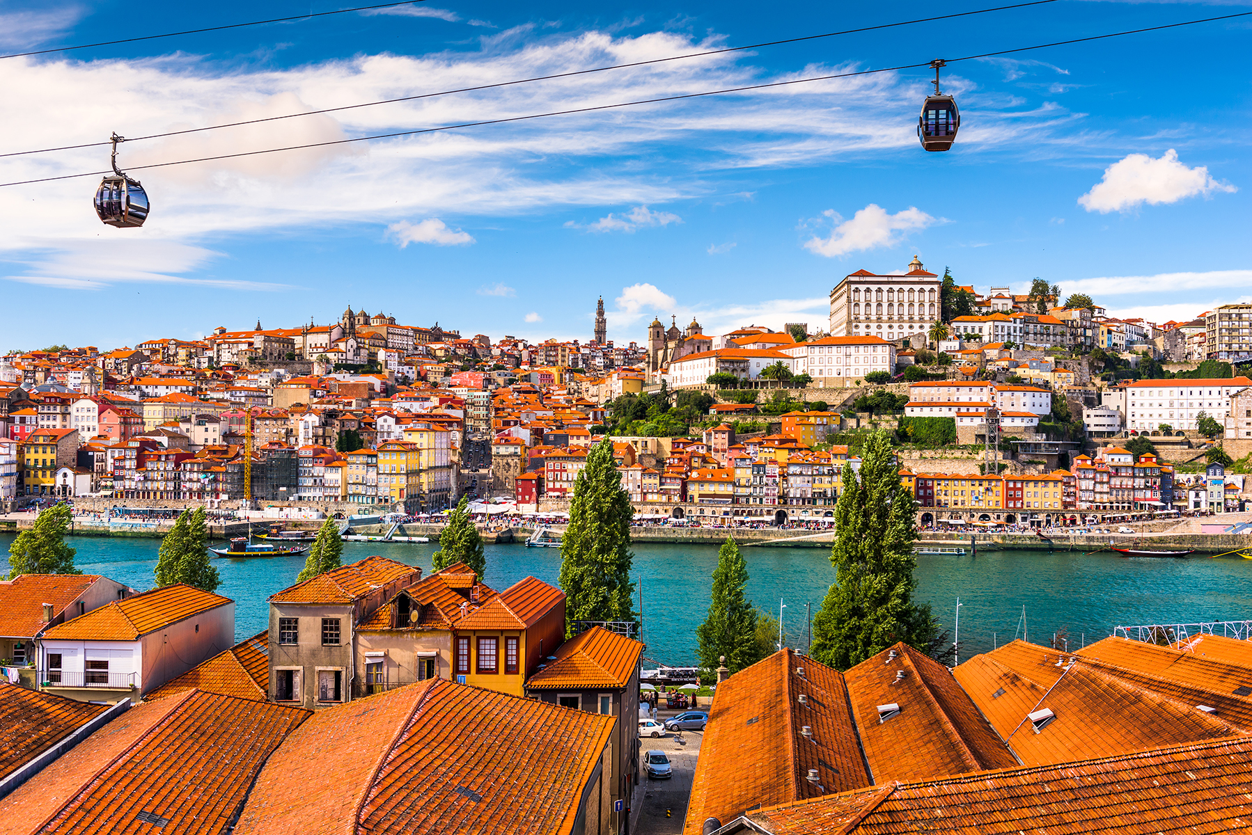 Clay rooftops in the foreground, a blue river in the middle ground, and beautiful houses of varying colors with clay roofs in the background. Two gondolas are in the sky,