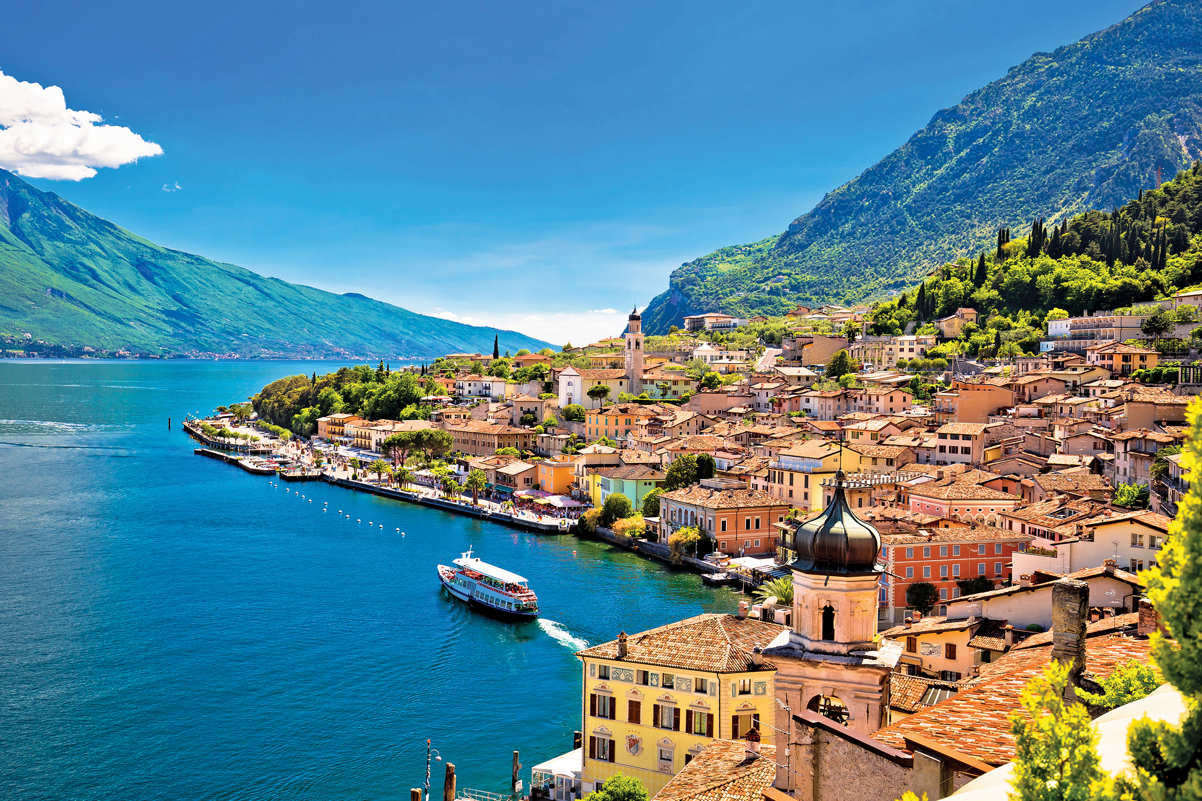 Lake Garda with a coastline filled with different color houses, mountains in the background, and a ferry boat in the middle. 