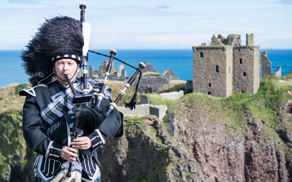 man playing the bag pipe with cliff in background